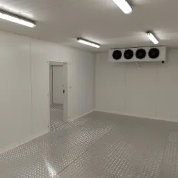 Wnętrze chłodni z panelową zabudową ścian i podłogą z blachy ryflowanej, widoczny agregat chłodniczy z czterema wentylatorami i oświetlenie jarzeniowe.