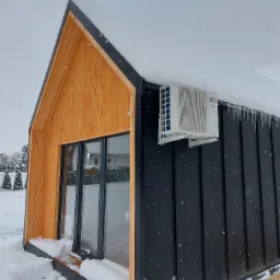 Nowoczesny domek z drewnianą fasadą i klimatyzatorem Cooper&Hunter na czarnej ścianie, pokryty śniegiem i zwisającymi soplami.