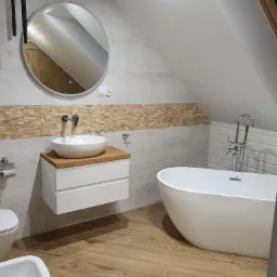 Nowoczesna łazienka na poddaszu z wolnostojącą wanną, umywalką nablatową na drewnianym blacie, bidetem i toaletą, wykończona płytkami imitującymi drewno i kamień.