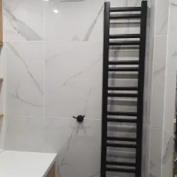 Biała łazienka z marmurowymi płytkami, czarnym grzejnikiem drabinkowym i wentylacją kratką u góry.