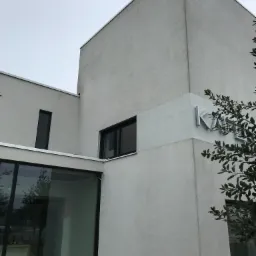 Nowoczesny budynek z białego tynku z dużymi oknami i minimalistycznym logo firmy na elewacji, widoczne fragmenty zieleni.