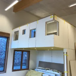 Wnętrze z antresolą w trakcie budowy, ściany z płyt kartonowo-gipsowych zabezpieczone taśmą, widoczne okna z widokiem na ceglany mur, sufit z oświetleniem punktowym.