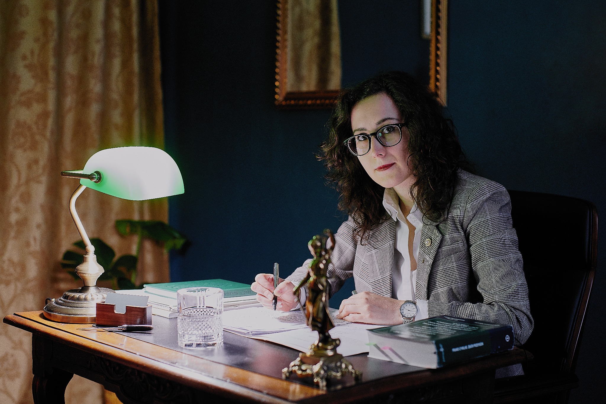 Kobieta w okularach i marynarce siedzi przy biurku z zieloną lampką, pisząc na papierze obok statuetki Temidy i książki 'Kodeks postępowania cywilnego'.