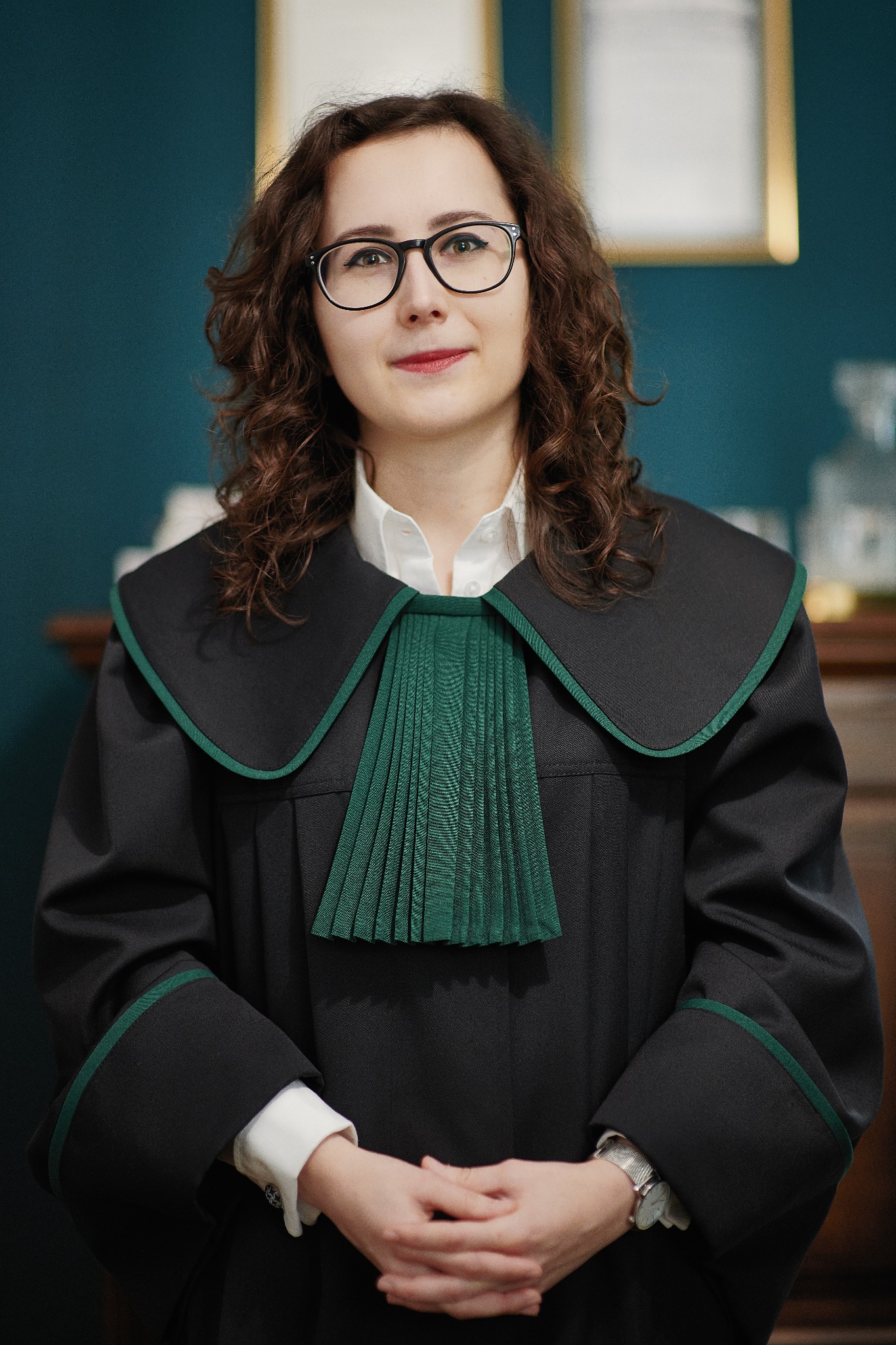 Portret kobiety w todze prawniczej z zielonym żabotem, splecione dłonie, okulary w czarnych oprawkach, brązowe kręcone włosy, turkusowe tło.