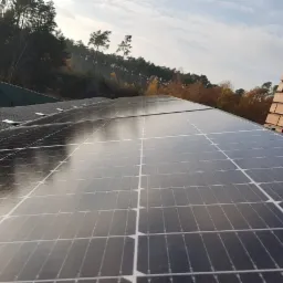 Panele słoneczne na dachu domu z kominem z cegły, widok z góry, w tle drzewa i błękitne niebo.