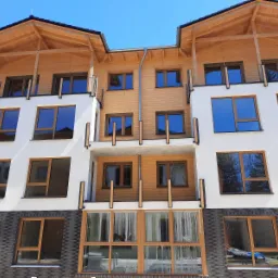 Nowoczesny budynek wielorodzinny z elementami drewnianymi, białymi ścianami i szarą cegłą na dole, widoczne okna z drewnianą ramą i balkony z drewnianymi balustradami, błękitne niebo w tle.