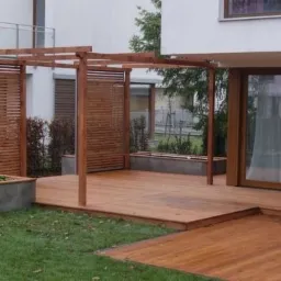 Drewniana pergola z ażurowymi ściankami na tarasie z desek, betonowe donice z zielenią, nowoczesny dom w tle.