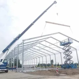 Montaż stalowej konstrukcji hali: żuraw podnosi element, a podnośnik koszowy służy do mocowania połączeń.