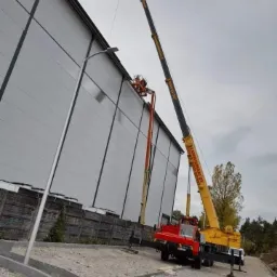 Żółto-czarny dźwig z wysięgnikiem i czerwonym podwoziem oraz pomarańczowy podnośnik koszowy na tle szarej hali przemysłowej. Na podnośniku widoczni pracownicy wykonujący prace na wysokości.