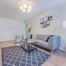 Jasne wnętrze salonu z szarą sofą, geometrycznym dywanem i lampą na trójnogu, minimalistyczny design.