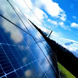 Nisko kątowe ujęcie paneli słonecznych odbijających błękitne niebo z chmurami na tle zalesionego zbocza.