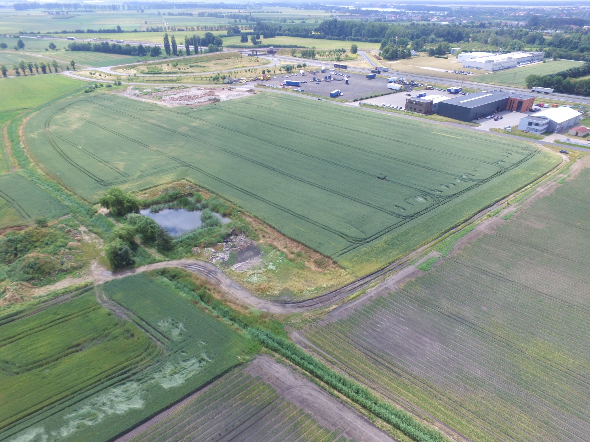 Panoramiczny widok z lotu ptaka na pola uprawne, mały staw, parking dla ciężarówek i budynki magazynowe w tle.