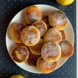 Kolekcja okrągłych placuszków posypanych cukrem pudrem na białym talerzu, obok dwa żółte jabłka na ciemnym tle w gwiazdki.