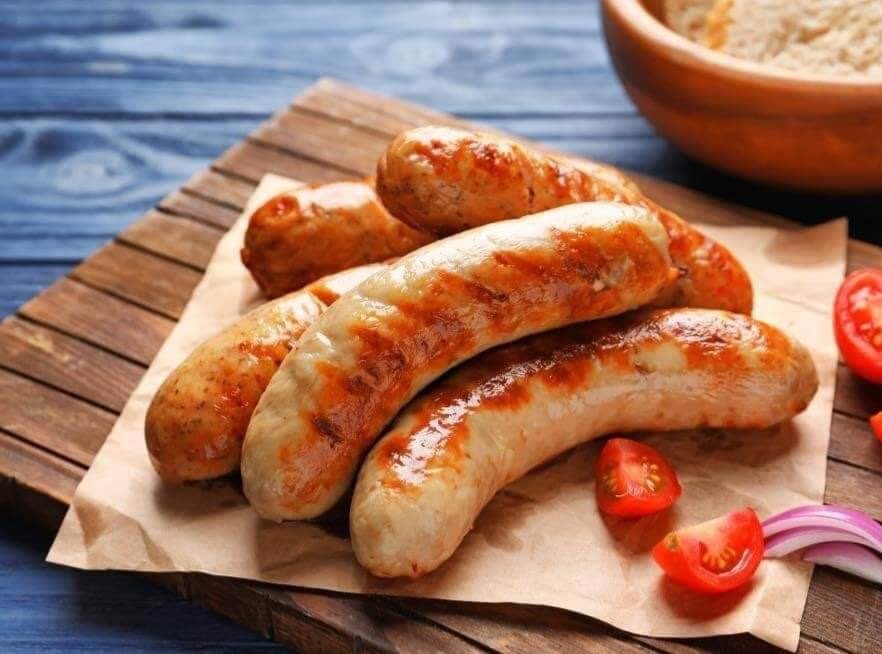 Grillowane kiełbaski z rumianymi przypaleniami, podane na papierze kraft na drewnianej desce z dodatkiem pomidorów i czerwonej cebuli, na tle niebieskich desek.