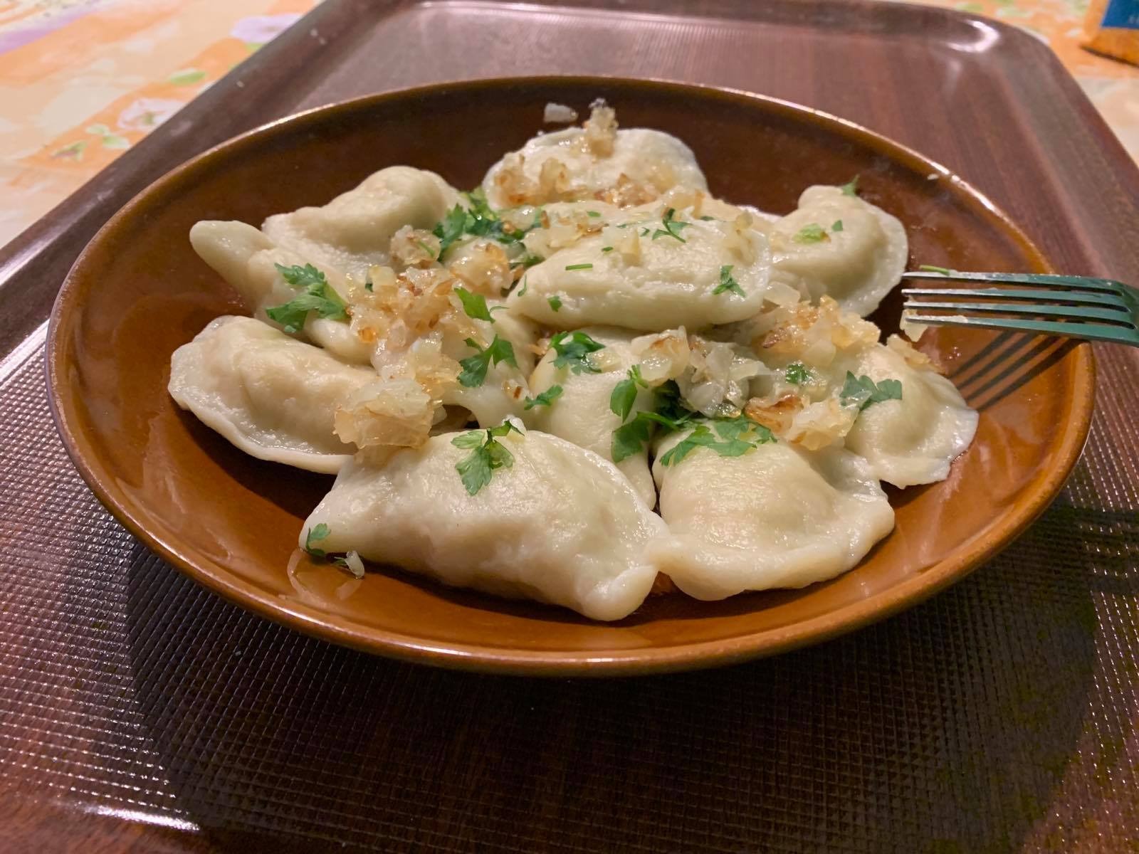 Brunatna misa z pierogami okraszonymi zeszkloną cebulką i posiekaną zieloną pietruszką, na tacy z widelcem wbijającym się w jednego z pierogów.