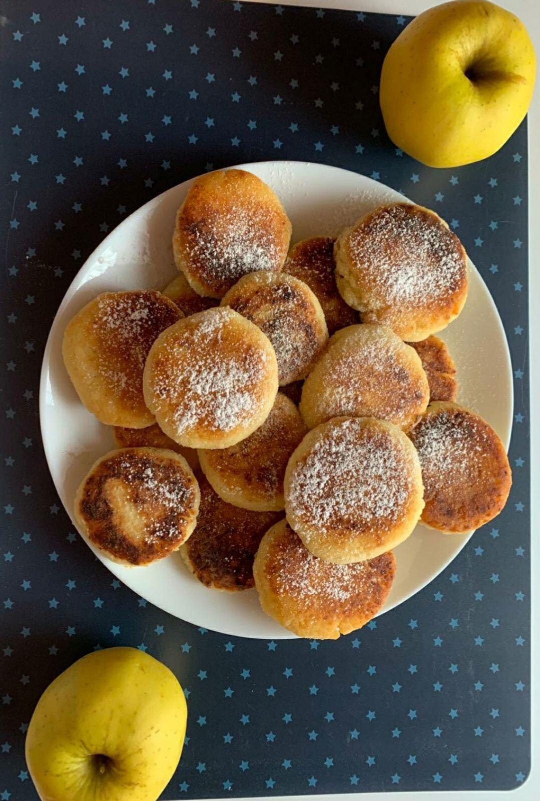 Kolekcja okrągłych placuszków posypanych cukrem pudrem na białym talerzu, obok dwa żółte jabłka na ciemnym tle w gwiazdki.