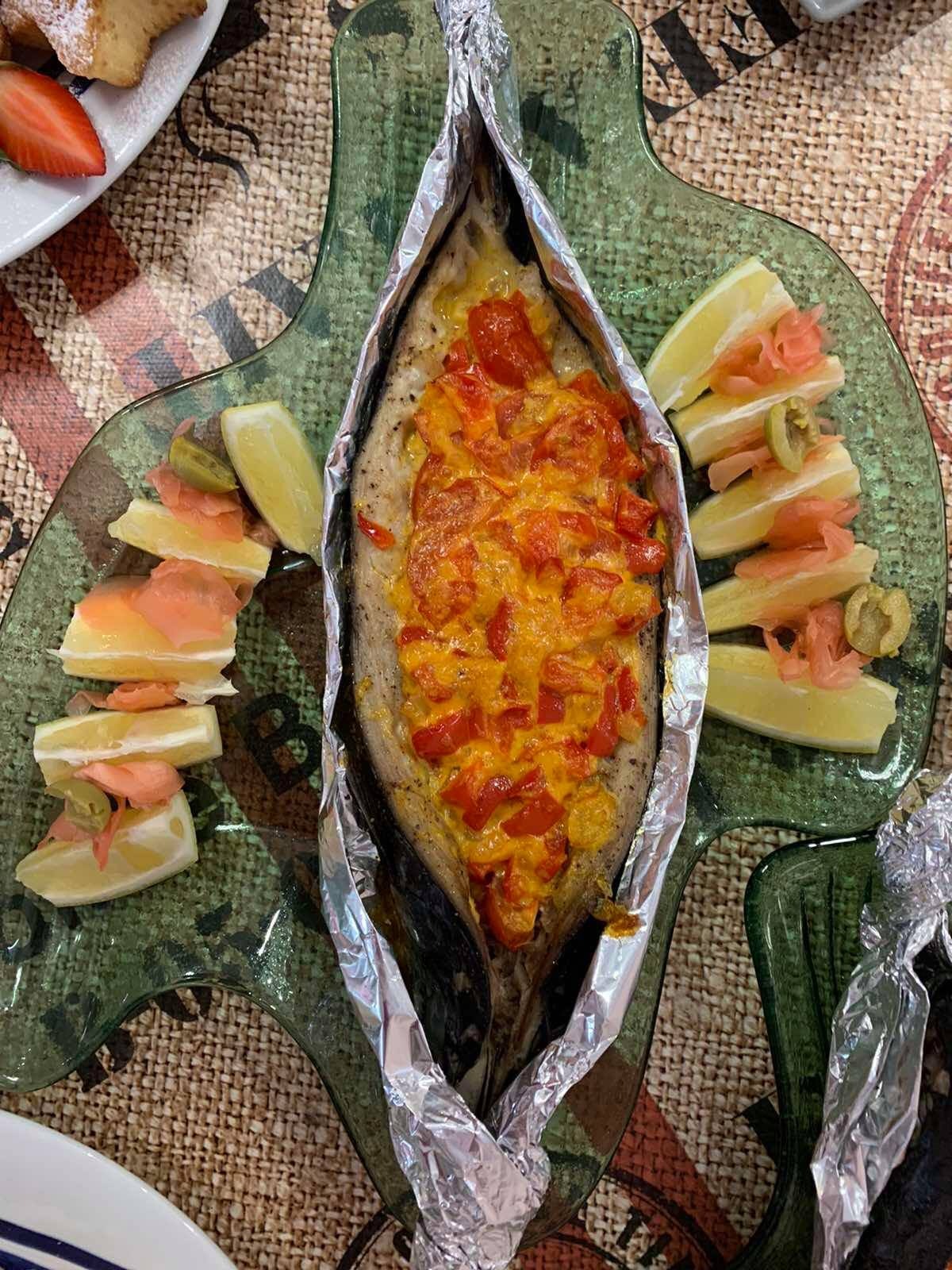 Upieczona ryba w folii aluminiowej, udekorowana czerwoną papryką i żółtym sosem, podana na zielonym talerzu z ćwiartkami cytryny i marynowanym imbirem.