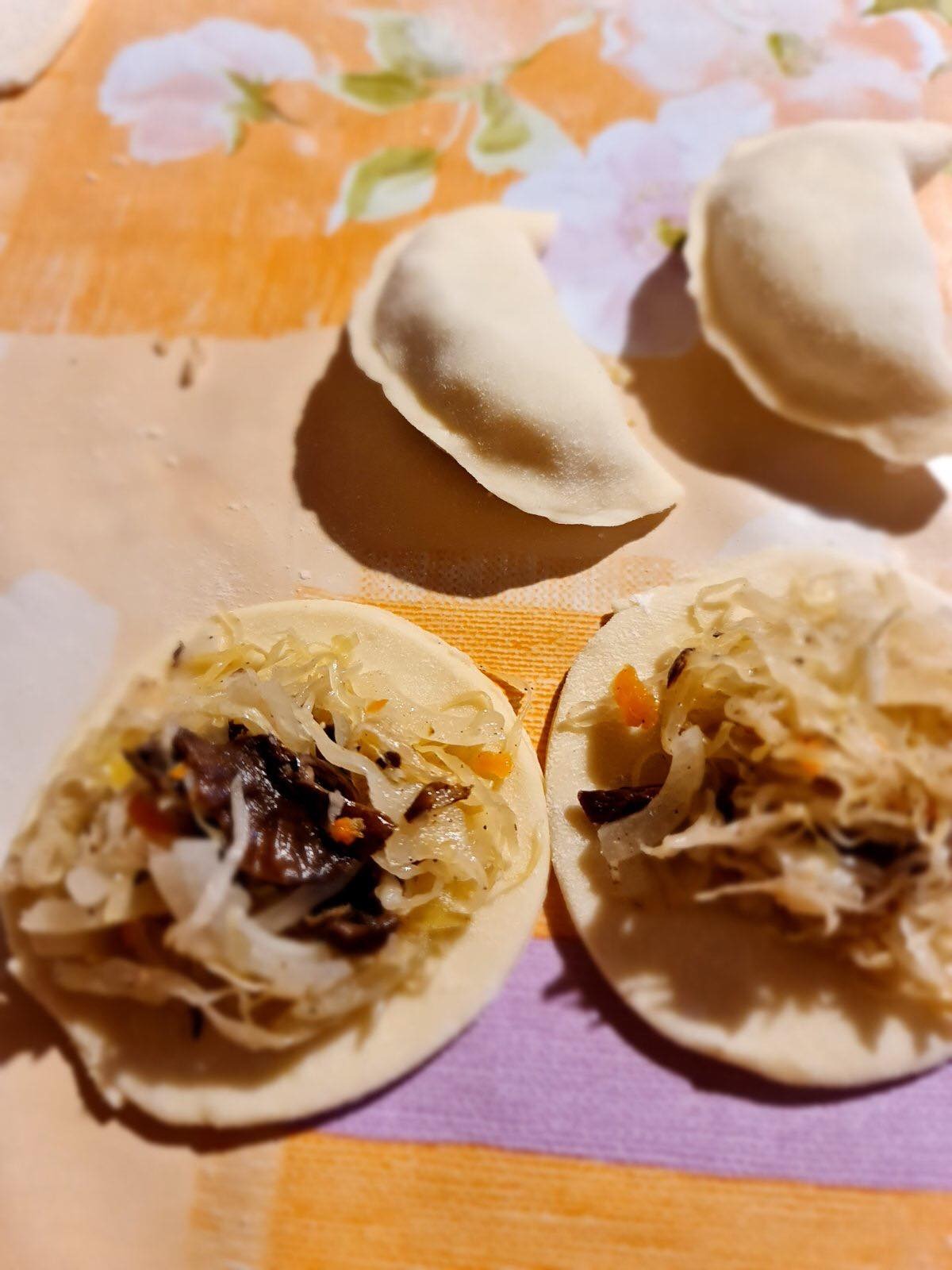 Przygotowywanie pierogów z kapustą i grzybami: dwa pierogi z farszem na surowym cieście, obok trzy uformowane, gotowe do gotowania, na wzorzystym tle.