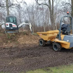 Żółta wywrotka Terex i biała minikoparka Bobcat E26 na placu budowy, widoczne ślady opon w błocie i operator w wywrotce.