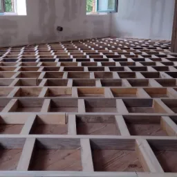 Drewniana konstrukcja legarów pod podłogę w trakcie remontu, widoczne stare deski pod spodem, surowe ściany w tle i pionowy słup podtrzymujący strop.