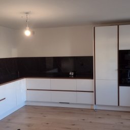 REMONTRY - Nowoczesna kuchnia z białymi szafkami, czarnym blatem i marmurowym backsplashiem. Widoczne oświetlenie i drewniana podłoga. Minimalistyczny design.