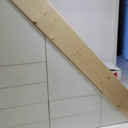 REMONTRY - Szafki z szufladami pod schodami, wykończone drewnianą belką. Białe fronty szuflad z delikatnymi poziomymi liniami. Widoczny fragment podłogi i ściany.