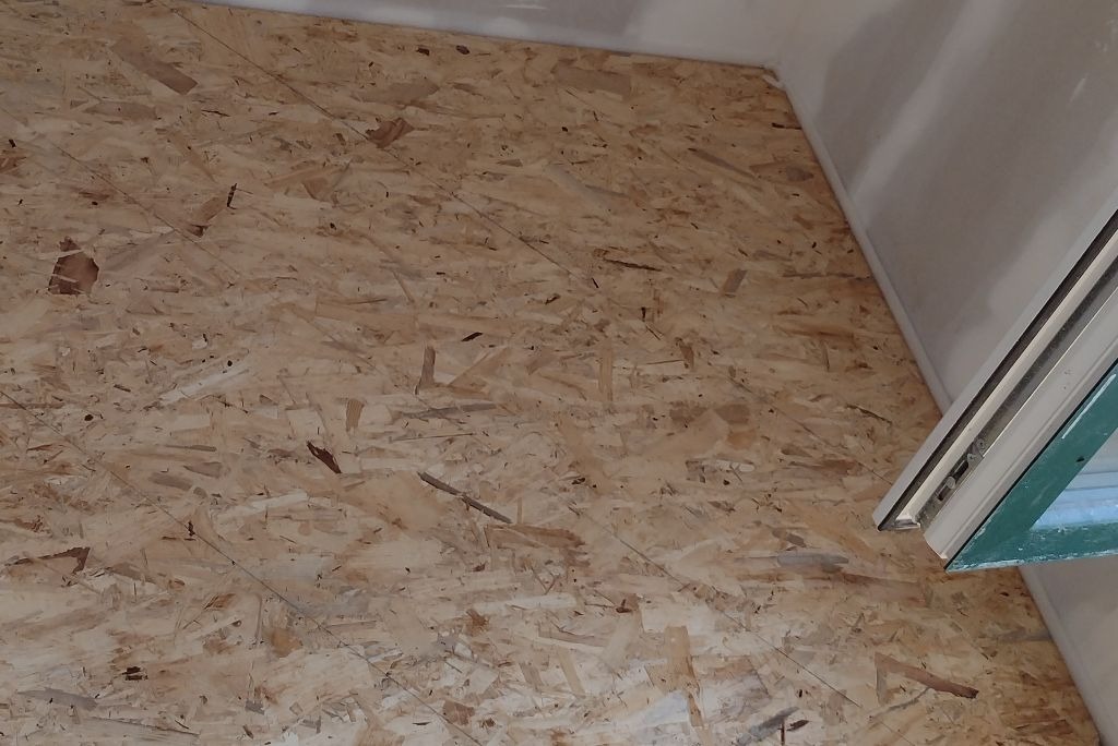 Surowa płyta OSB na podłodze, widoczny fragment okna i ściany w rogu pomieszczenia.