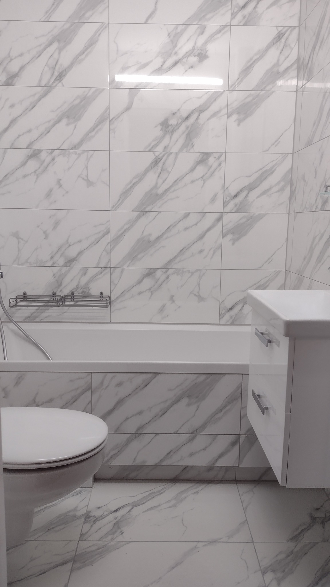 Wykończona łazienka z białymi płytkami imitującymi marmur na ścianach i podłodze, widoczna toaleta, wanna z parawanem i biała szafka z szufladami.