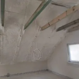 Poddasze po ociepleniu, widoczne drewniane belki stropowe i zwisający kabel elektryczny, okno zabezpieczone folią.