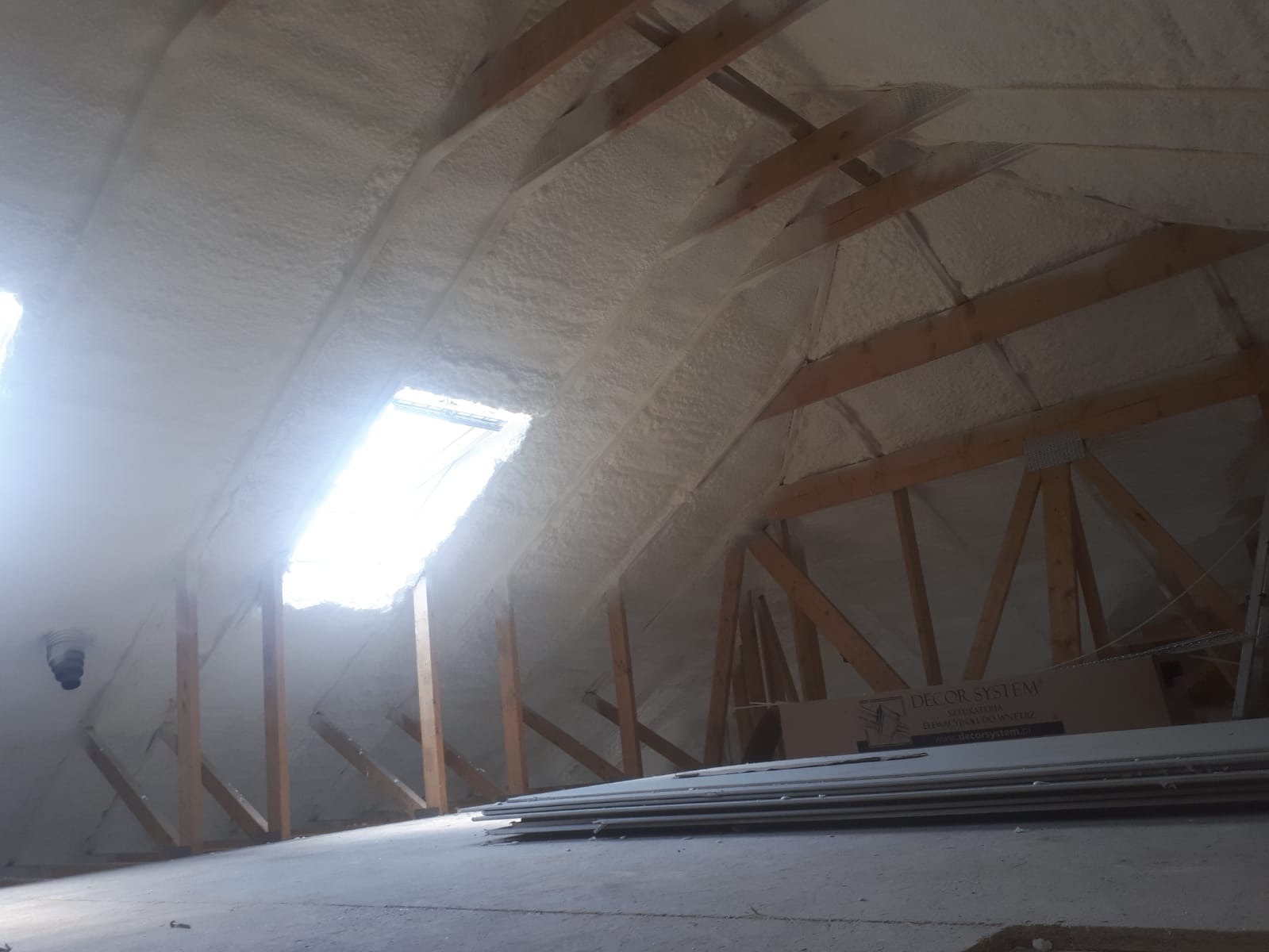 Poddasze po natrysku izolacji, widoczne krokwie i okno dachowe z silnym światłem słonecznym, na podłodze płyty gipsowe.