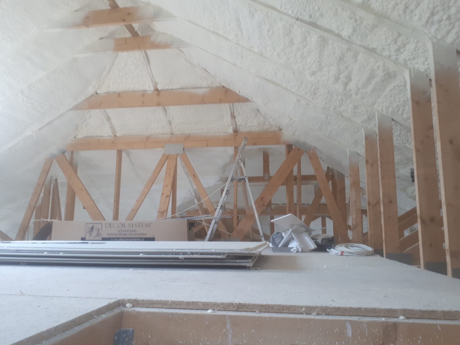Poddasze z drewnianą konstrukcją dachu pokryte białą warstwą izolacyjną, widoczna drabina i materiały budowlane.