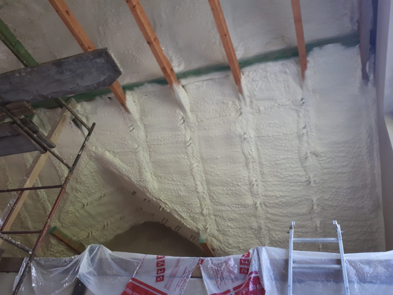 Poddasze ocieplone pianką PUR, widoczne drewniane belki konstrukcyjne, fragment rusztowania i drabiny, zabezpieczona folią przestrzeń poddasza.