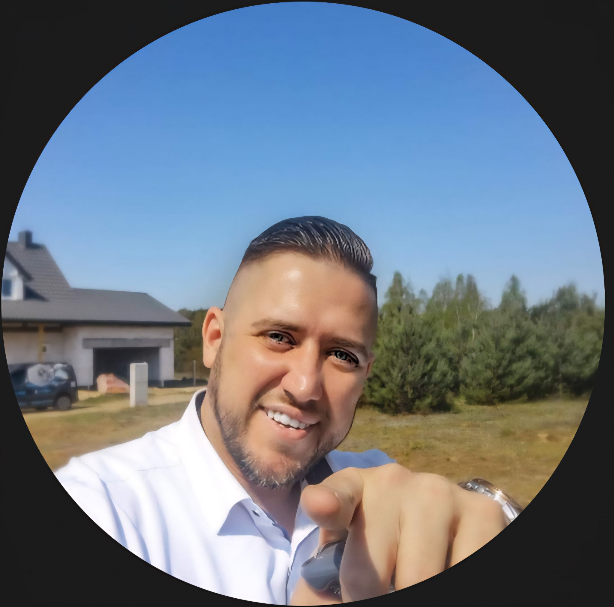 Uśmiechnięty mężczyzna w białej koszuli wskazuje palcem na tle domu w trakcie budowy i błękitnego nieba. Widoczny samochód. Kadr w okręgu.