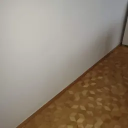 Ściana pomalowana na biało sąsiaduje z ułożoną parkietową podłogą o wzorze 3D, listwa przypodłogowa z drewna.