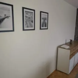 Świeżo pomalowana ściana w Pruszkowie z trzema czarno-białymi obrazami w ramkach przedstawiającymi molo, komoda z drewnianym blatem i parkiet na podłodze.