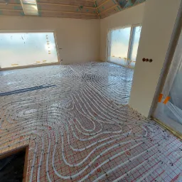 Białe rury ogrzewania podłogowego ułożone spiralnie na czerwonej siatce w jasnym pomieszczeniu w trakcie budowy, widoczne okno dachowe i okna zabezpieczone folią.