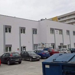 Jma budownictwo - Dwupiętrowy budynek biurowy z szarą elewacją i białymi ramami okiennymi, widoczne zaparkowane samochody i kontener na śmieci na pierwszym planie, pracownik na rusztowaniu przy ścianie budynku.