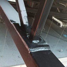 Jma budownictwo - Spojrzenie z dołu na fragment konstrukcji stalowej budynku, gdzie elementy pionowe łączą się z poziomymi za pomocą spawów i śrub; widoczne ślady świeżego malowania w kolorze czarnym i szarym.