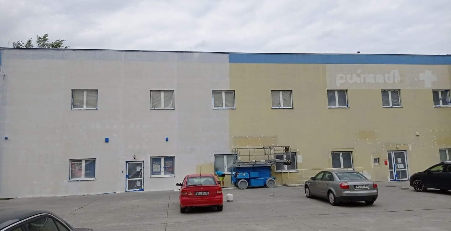 Malowanie elewacji budynku biurowego w trakcie realizacji, widoczny podnośnik koszowy, częściowo pomalowana ściana, okna zabezpieczone folią, zaparkowane samochody.