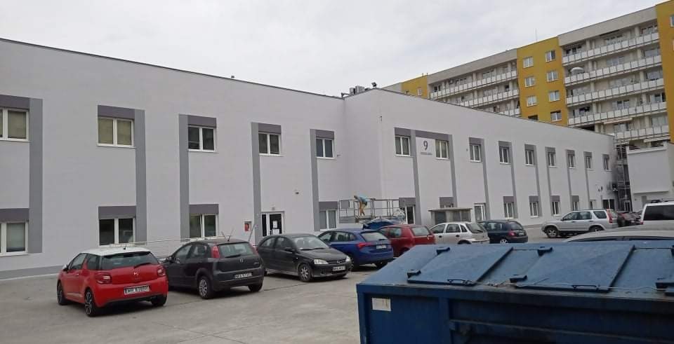 Dwupiętrowy budynek biurowy z szarą elewacją i białymi ramami okiennymi, widoczne zaparkowane samochody i kontener na śmieci na pierwszym planie, pracownik na rusztowaniu przy ścianie budynku.