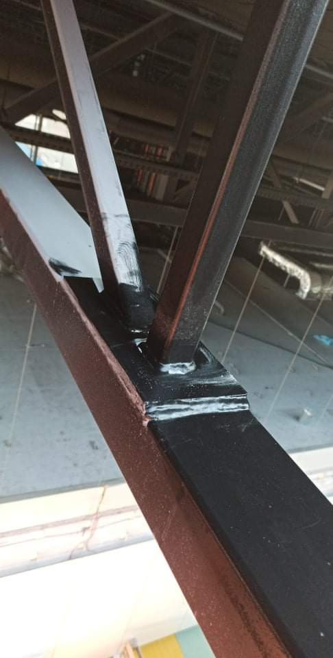 Spojrzenie z dołu na fragment konstrukcji stalowej budynku, gdzie elementy pionowe łączą się z poziomymi za pomocą spawów i śrub; widoczne ślady świeżego malowania w kolorze czarnym i szarym.