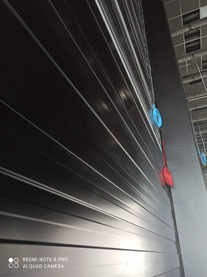 Zamknięta, czarna brama rolowana w hali przemysłowej, widoczne zwisające, kolorowe kable elektryczne.