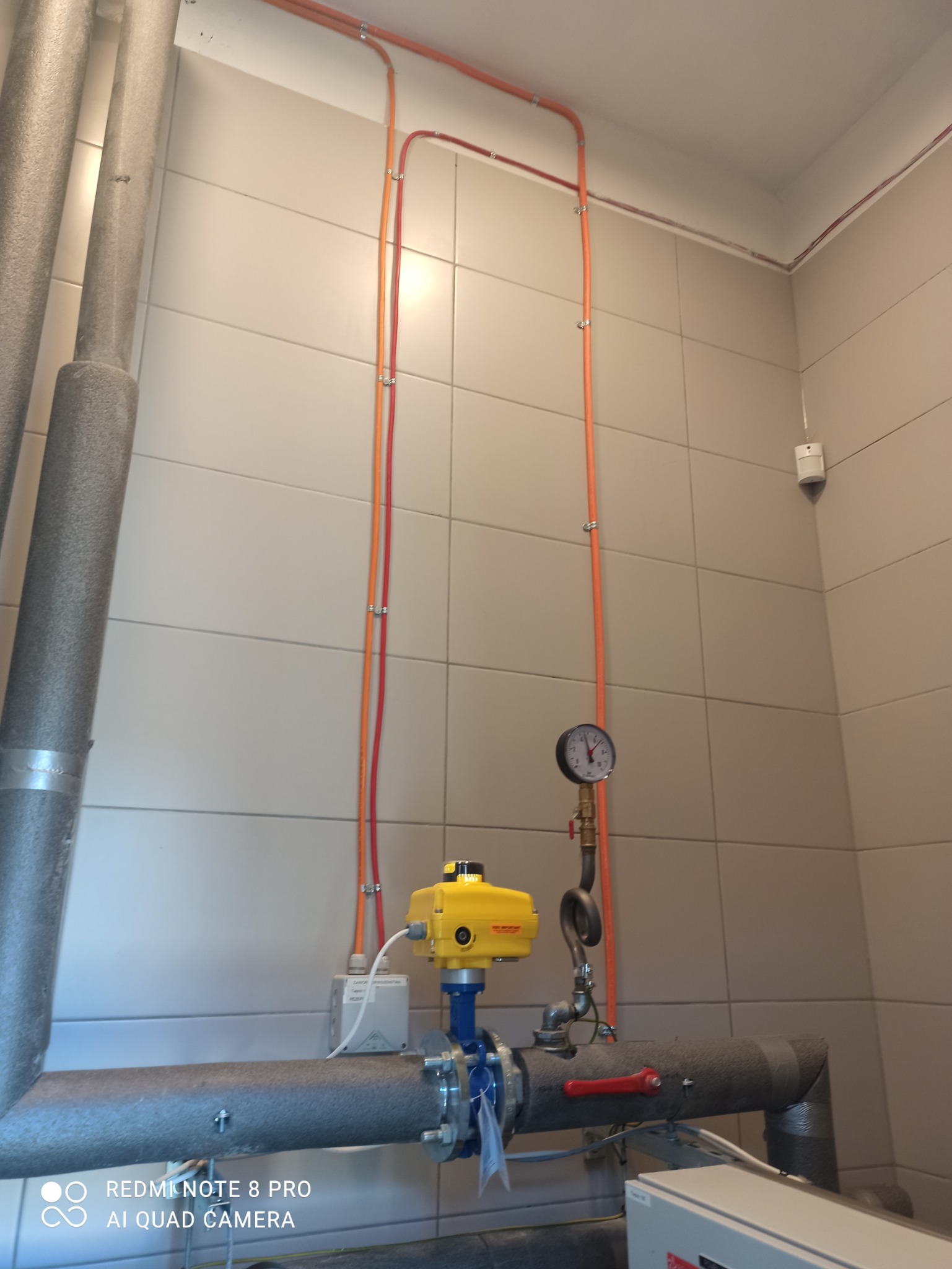 Instalacja grzewcza z rurami w izolacji, żółtym zaworem i manometrem na tle płytek ceramicznych.