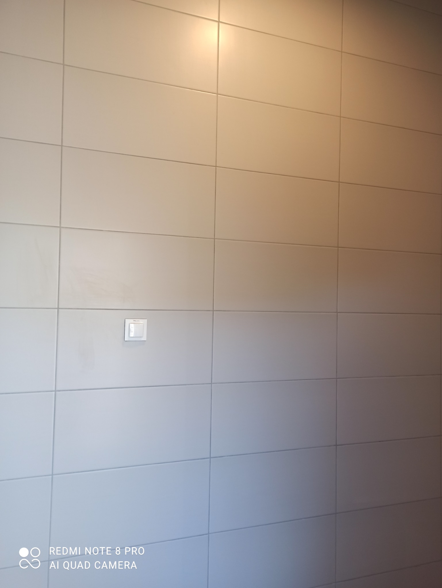 Ściana wyłożona jasnymi, kwadratowymi płytkami z widocznym włącznikiem światła