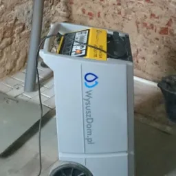 Profesjonalny osuszacz powietrza z logo firmy WyszuszDom.pl na tle ceglanej ściany i fragmentu posadzki.