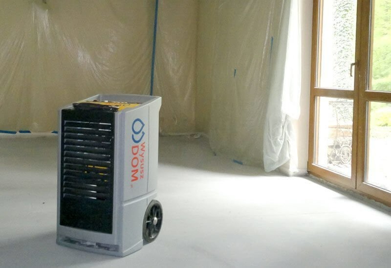 Przenośny osuszacz powietrza marki Wysusz Dom na kółkach, w pomieszczeniu zabezpieczonym folią malarską, widoczne okno z drewnianą ramą.