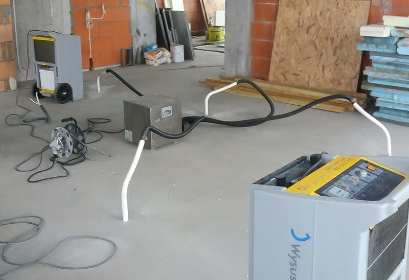 Dwa osuszacze powietrza marki Wysus.pl, połączone przewodami, stoją na szarym, betonowym podłożu w surowym wnętrzu budynku w trakcie prac remontowych.