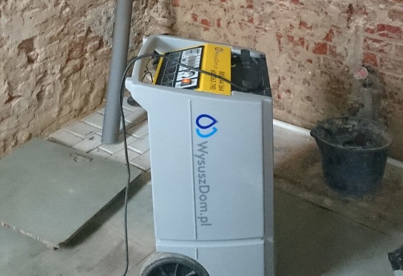 Profesjonalny osuszacz powietrza z logo firmy WyszuszDom.pl na tle ceglanej ściany i fragmentu posadzki.