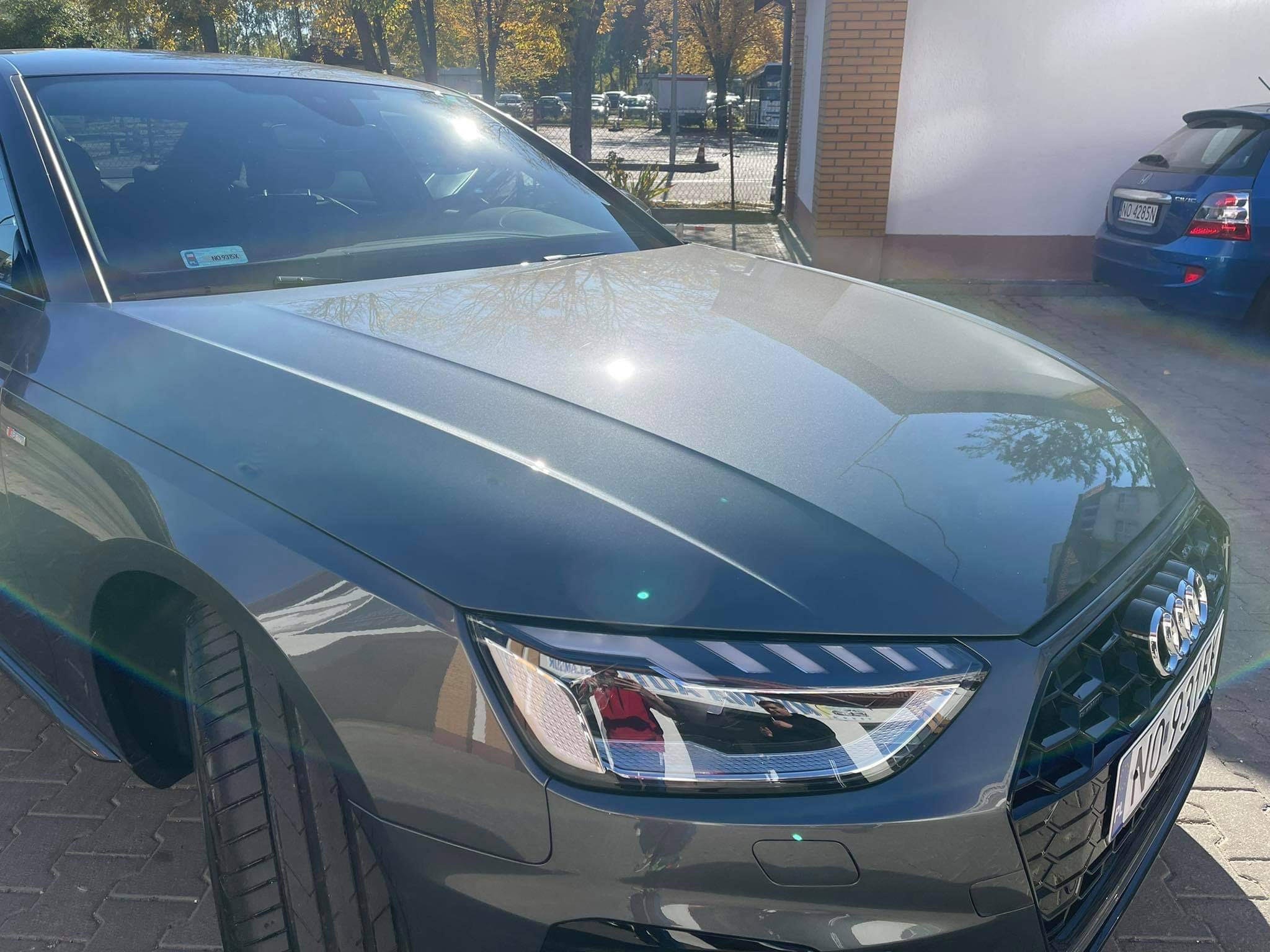 Błyszczący, grafitowy Audi A4 po umyciu, z widocznym odbiciem słońca na masce i przedniej szybie, zaparkowany na kostce brukowej przed budynkiem z cegły.