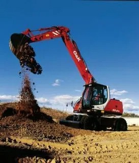 Czerwono-biała koparka kołowa marki O&K w trakcie pracy, wysypująca urobek na pryzmę ziemi na tle błękitnego nieba.